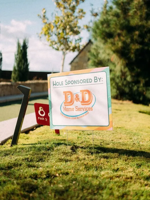 Golf course hole sponsor sign reading 'Hole Sponsored By: D&D Plumbing, Heating, and Air Conditioning' on a grassy green with a nearby tee marker.