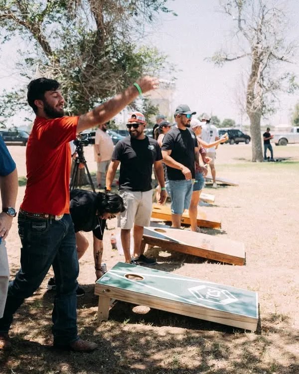 Group of people playing cornhole outdoors; man in a red shirt throws a bag toward a green board while others watch on a sunny day.
