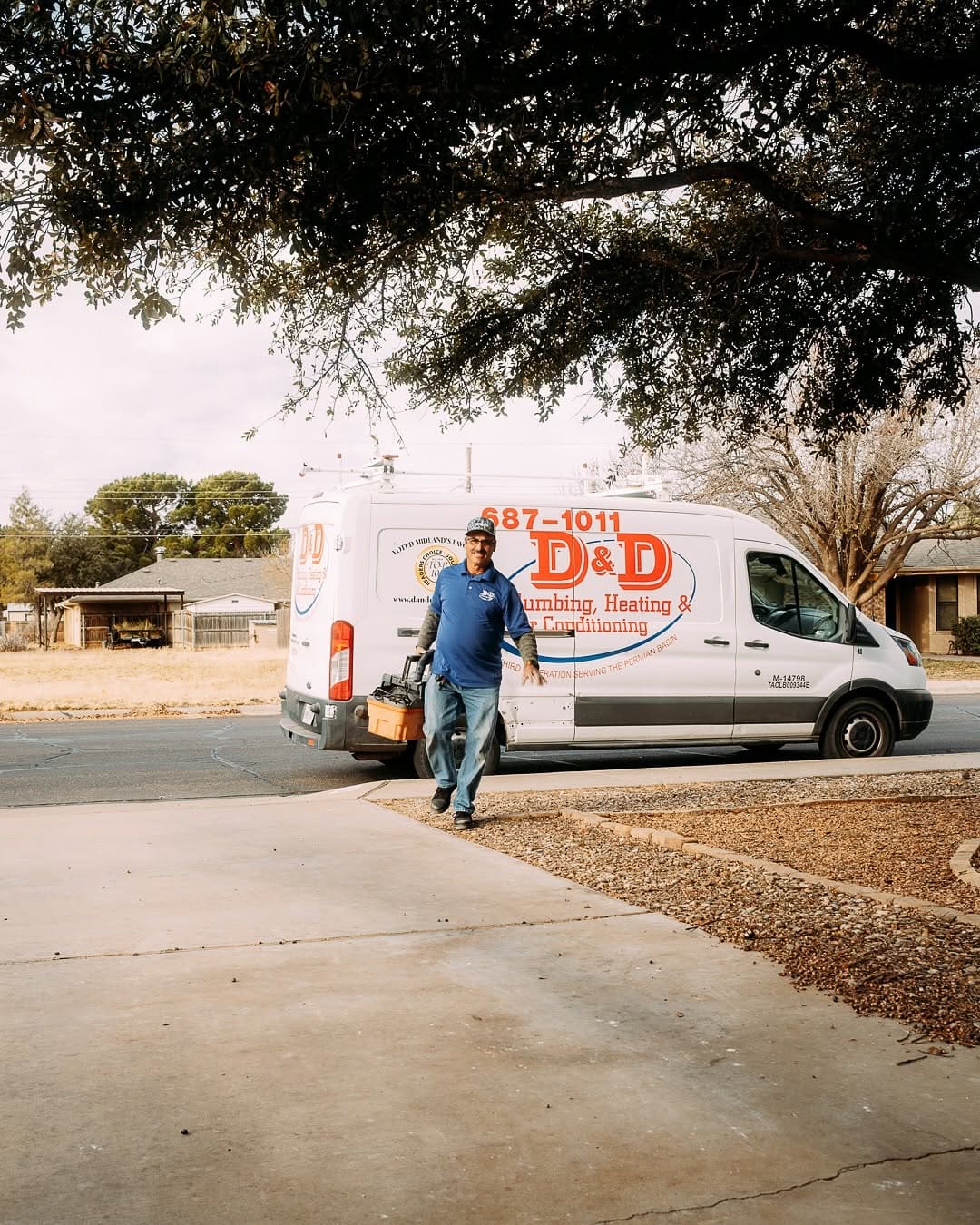 A tradesman in a blue shirt walks away from a white D&D plumbing van, carrying a toolbox on a sunny suburban street.