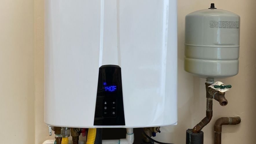 Tankless water heater installed inside a home with digital temperature display, water connections, gas line, and expansion tank.