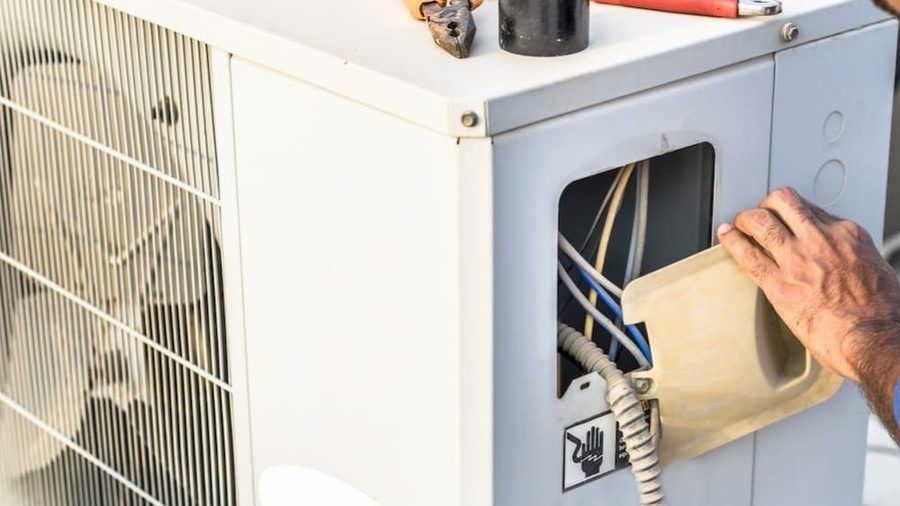 HVAC technician servicing the electrical components of an outdoor air conditioning condenser during a professional AC maintenance visit.